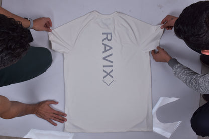 Two people folding white t-shirt with 'RAVIX' branding on a light gray surface.