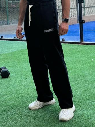 Model standing in black sweatpants on a sports court with a blue surface and green grass, wearing black athletic wear.